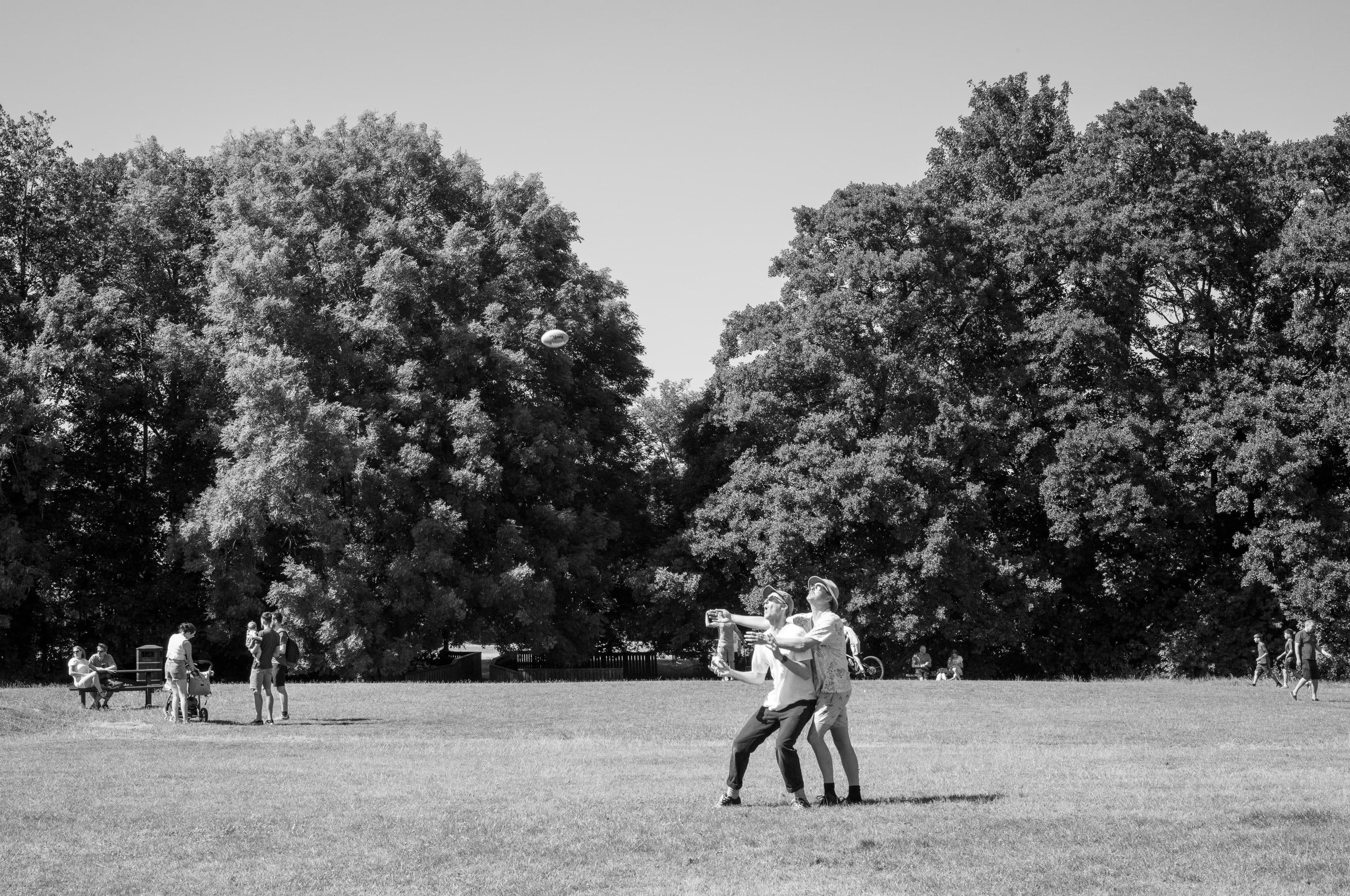 Two men in hats colliding as they both reach for a rugby ball in a park, trees behind