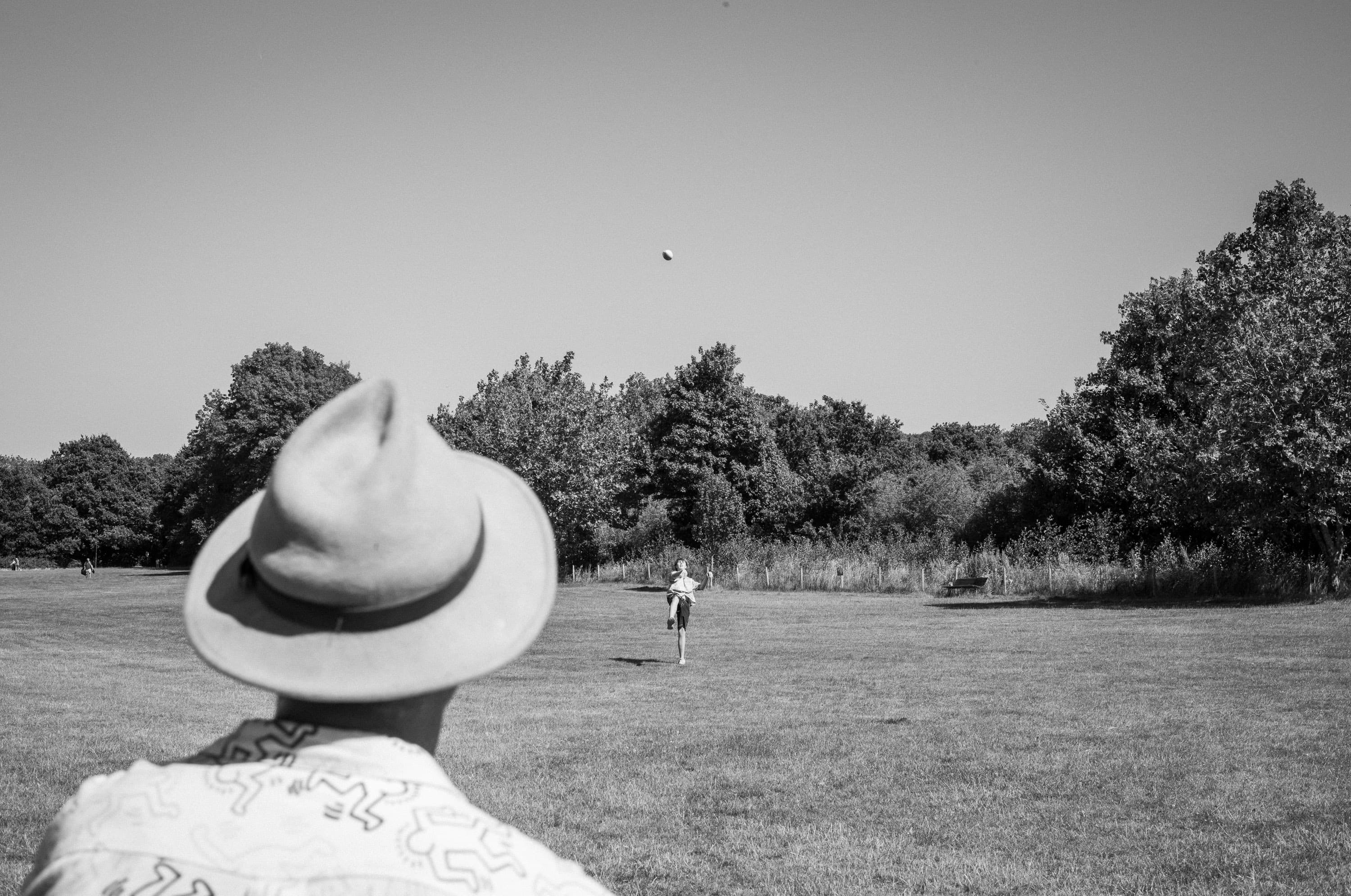 A man in a straw hat and a Keith Haring shirt, seen from behind, watching a ball arc high over a park while someone else runs to catch it