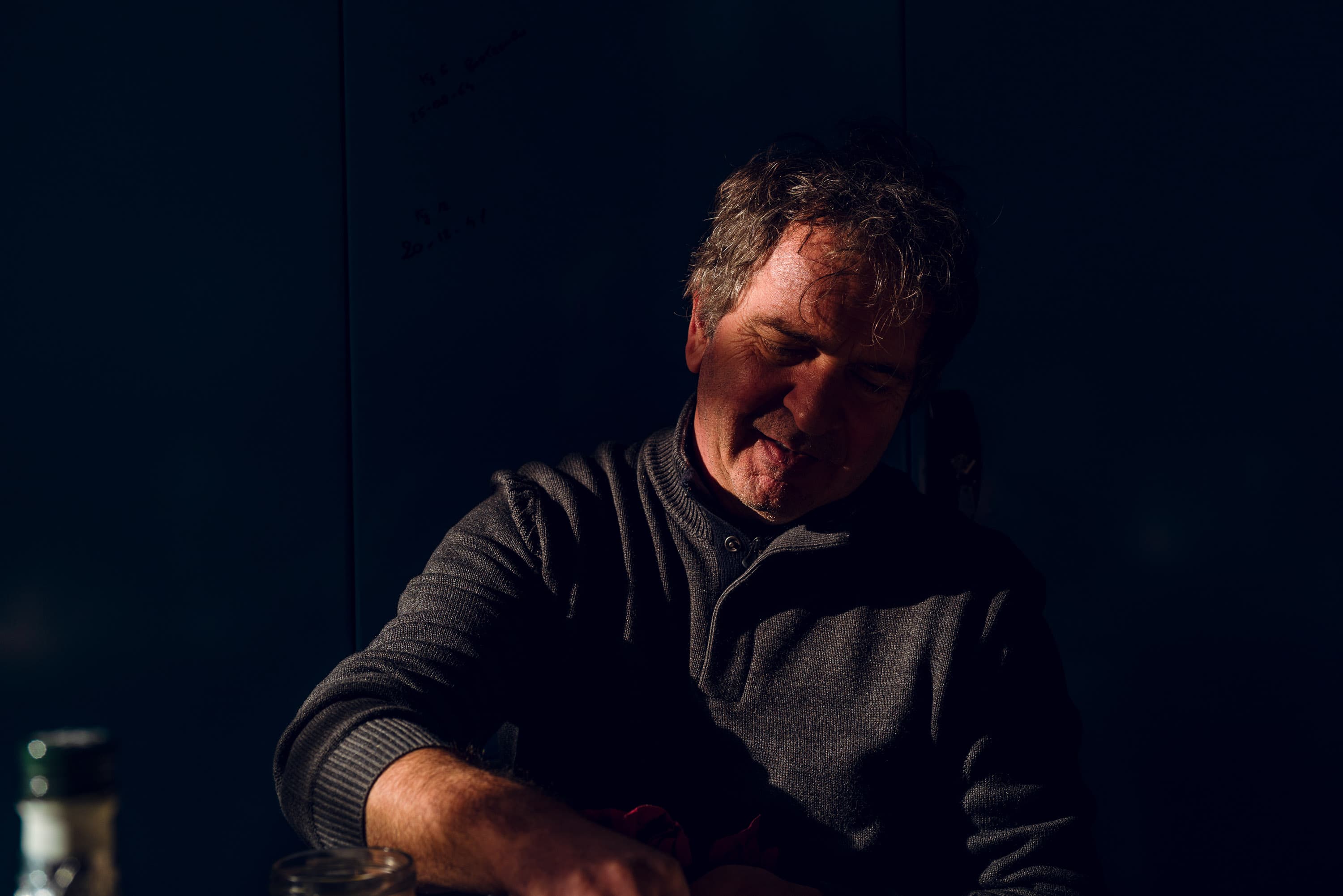 Pietro of Pegoretti sat at a table in a single pool of warm light against a dark wall, head down, in the middle of telling a story, a jar and a bottle at his elbow